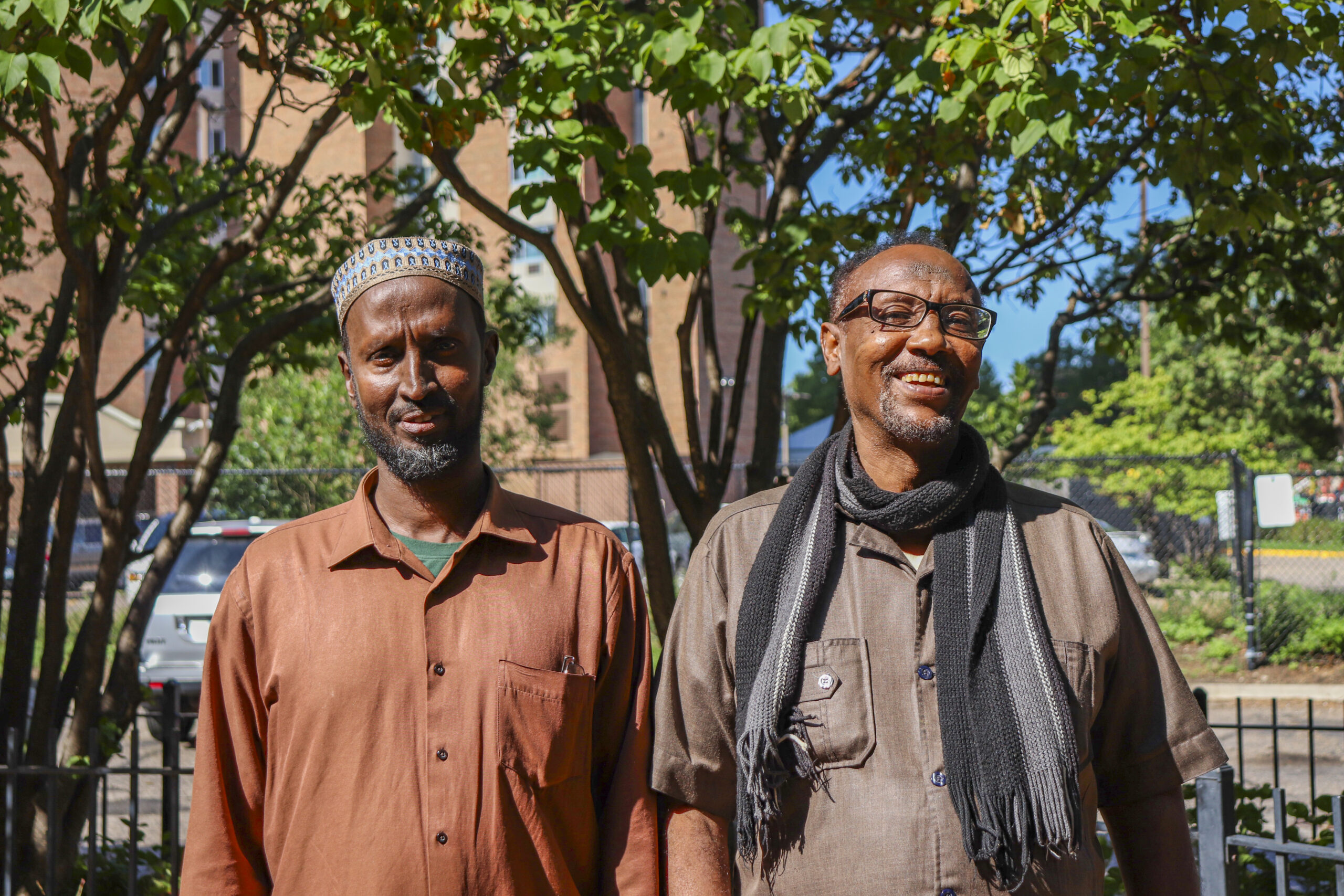 Aeon Affordable Homes Ahmed & Hassan stand outside, smiling, as their story unfolds amid trees and a sunlit building in the background.