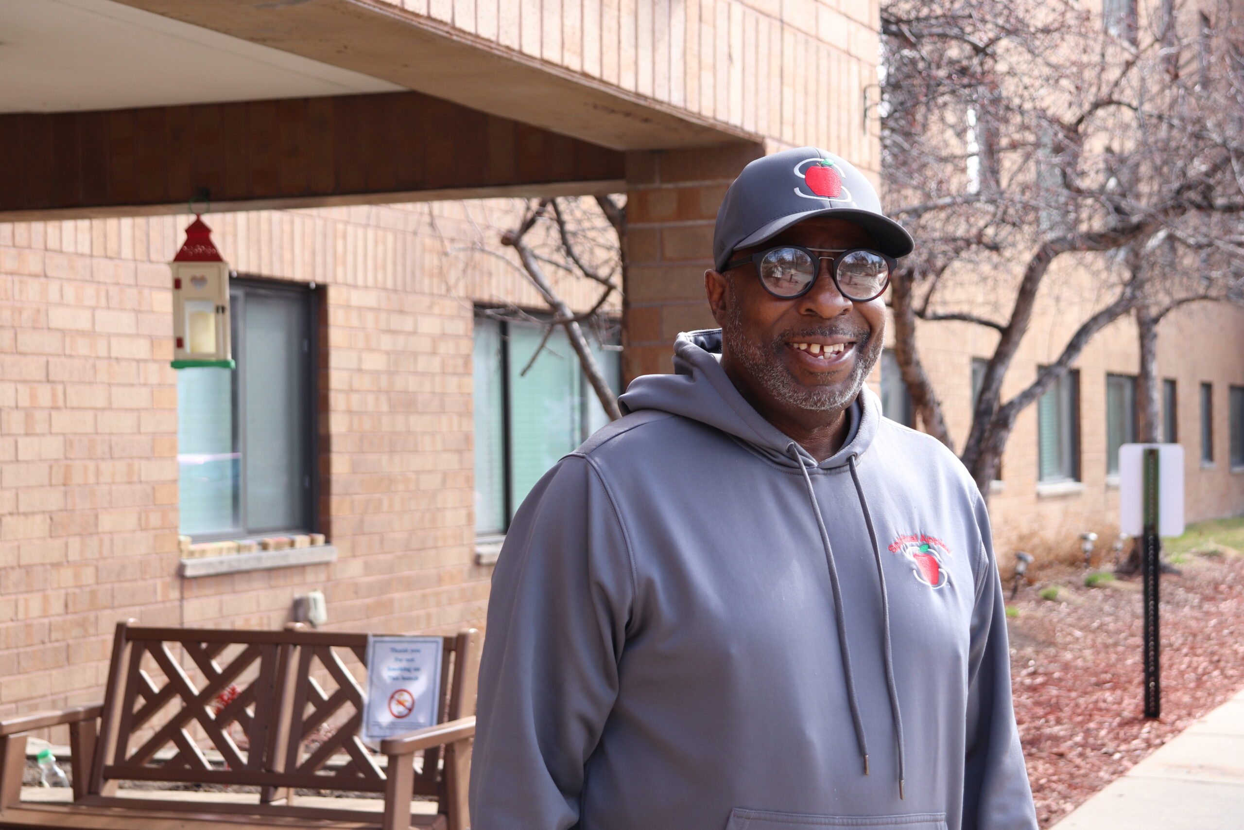 Aeon Affordable Homes Smiling man in a gray hat and hoodie stands outside a brick building, near a bench and bird feeder, sharing James's Story.