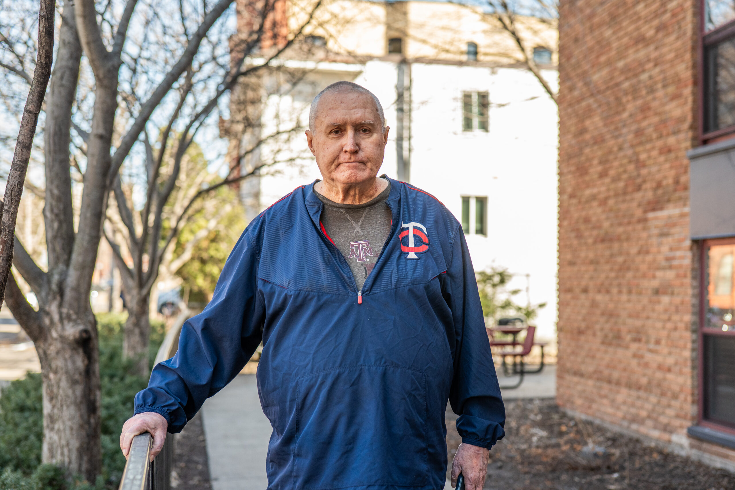 Aeon Affordable Homes An older man in a blue jacket stands outside on a sidewalk—perhaps it's part of Rick's Story—near brick buildings and leafless trees.