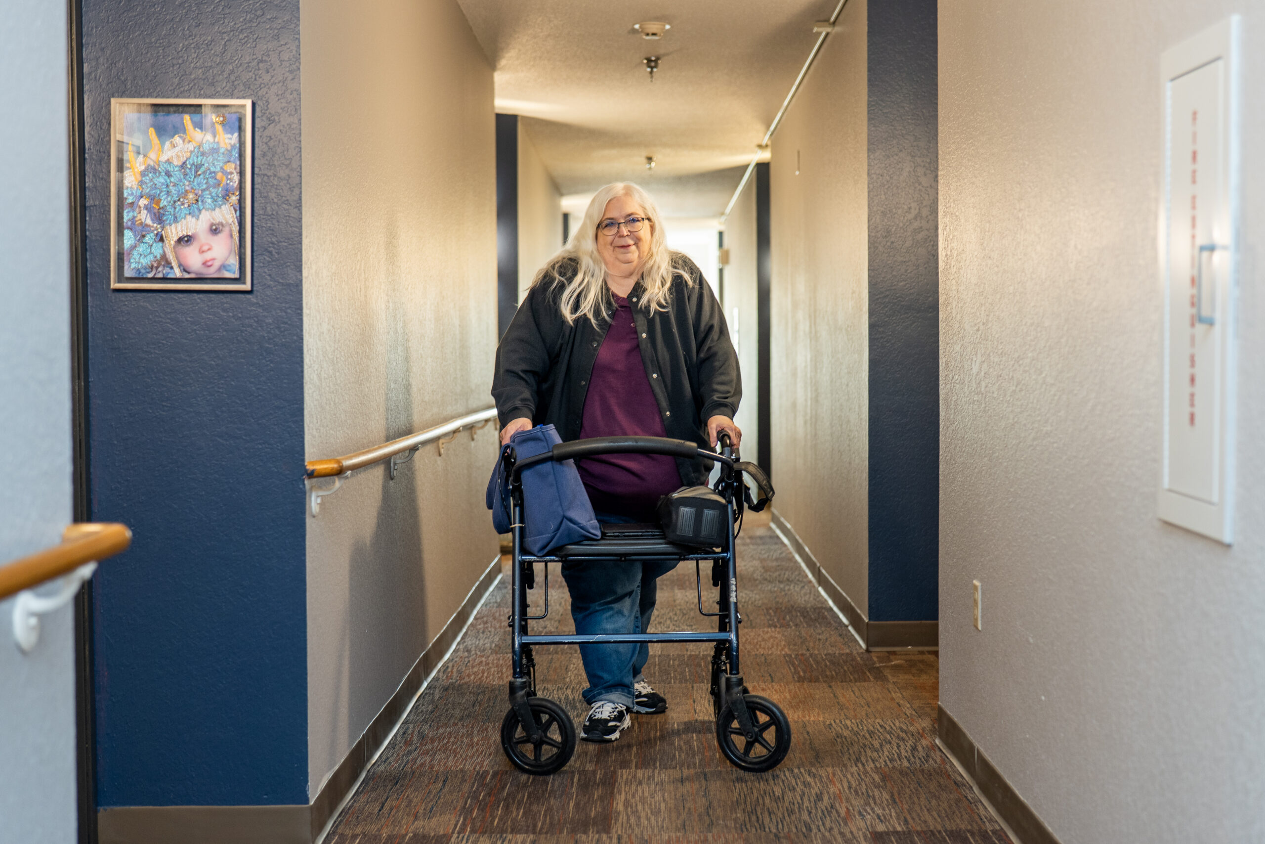 Aeon Affordable Homes An older woman with long white hair uses a walker in a hallway, smiling at the camera—this is part of Dianne's Story.