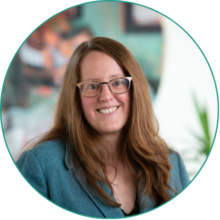 Aeon Affordable Homes Smiling woman, Caroline, with long brown hair and glasses in a blue blazer, Interim CEO, stands before a blurred background.