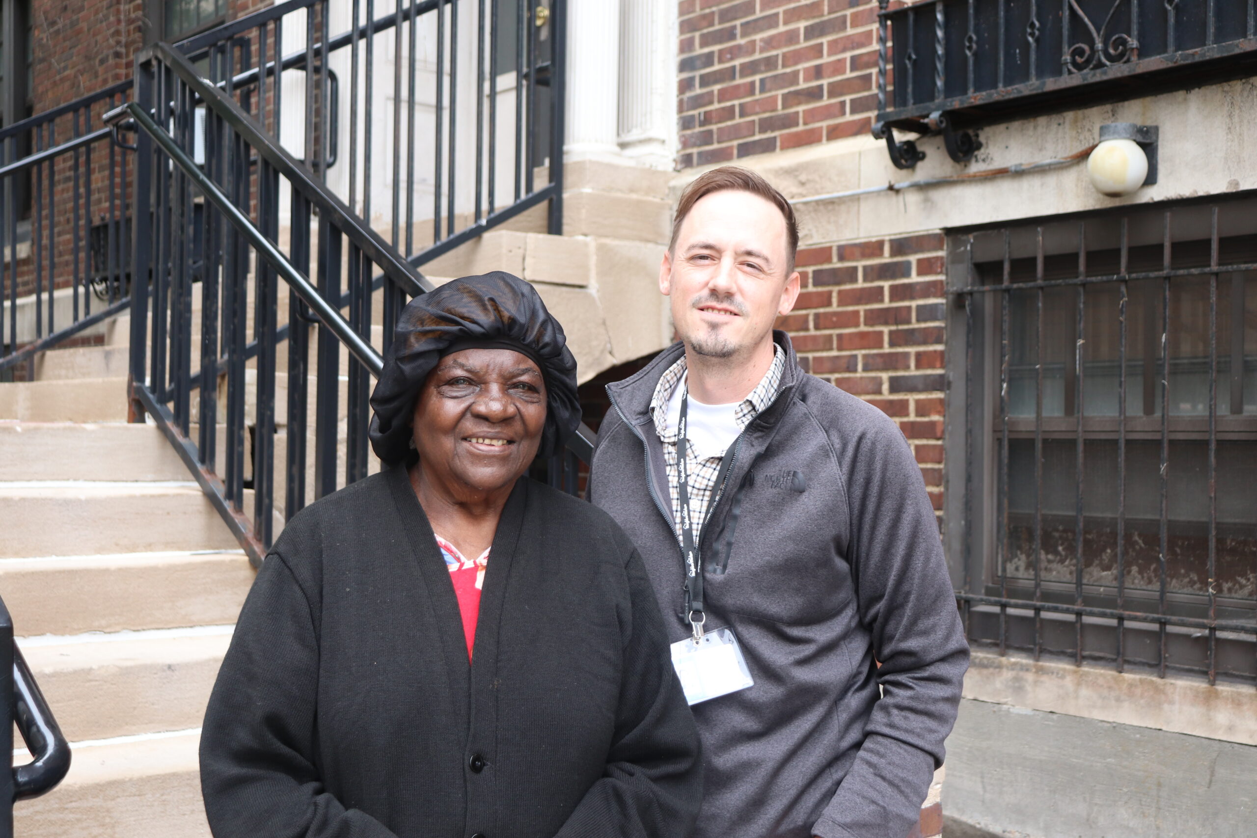 Aeon Affordable Homes Two people stand smiling together outside a brick building near a staircase.