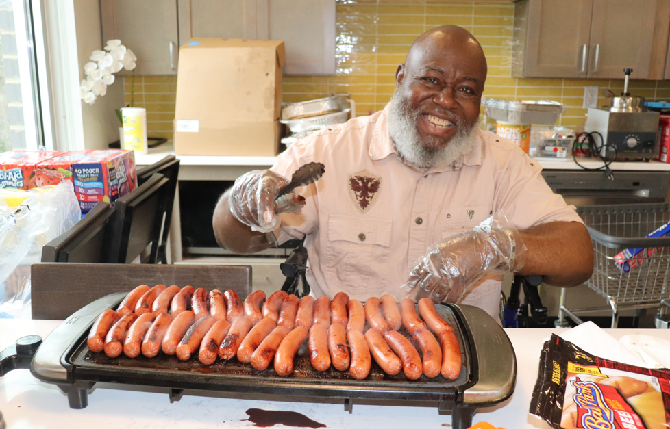 Aeon Affordable Homes Smiling man grilling hot dogs indoors, wearing gloves and holding tongs, ready to serve neighbors for National Night Out.