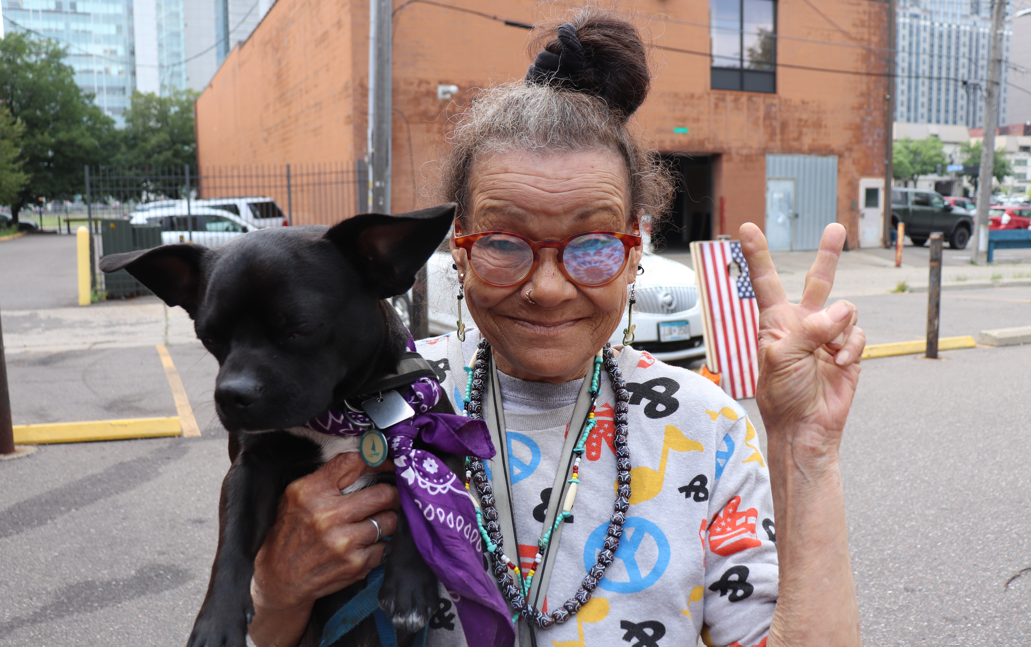 Aeon Affordable Homes Older woman with glasses holds a black dog and flashes a peace sign with an American flag at a National Night Out community event.