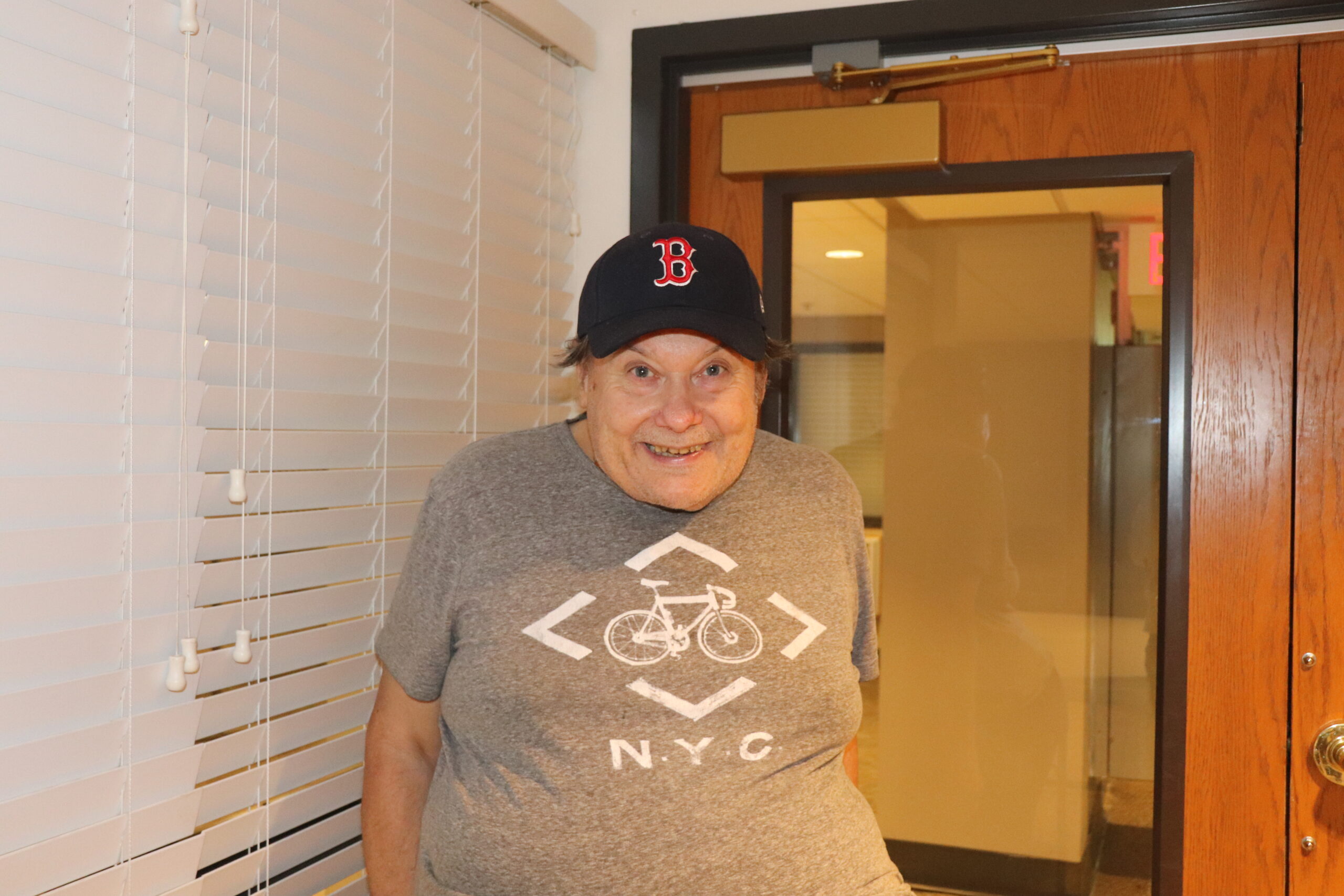 Aeon Affordable Homes Smiling elderly man in a Boston Red Sox hat and NYC bike t-shirt stands indoors by a door and window blinds.