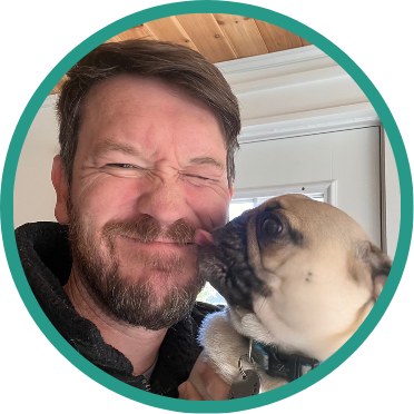 Aeon Affordable Homes Smiling man with a beard is being licked on the face by a small, light brown dog—Aeon staff always give back, even to their furry friends.