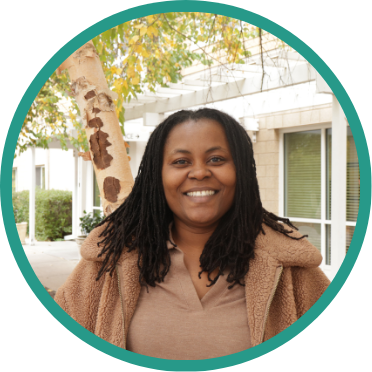 Aeon Affordable Homes Smiling woman with long locs stands outside near a tree, wearing a tan jacket and brown shirt, embodying the spirit of giving back.