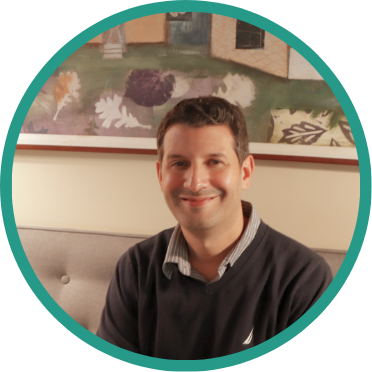 Aeon Affordable Homes A man with short brown hair sits on a couch, smiling, giving back to his community, with a painting hanging on the wall behind him.