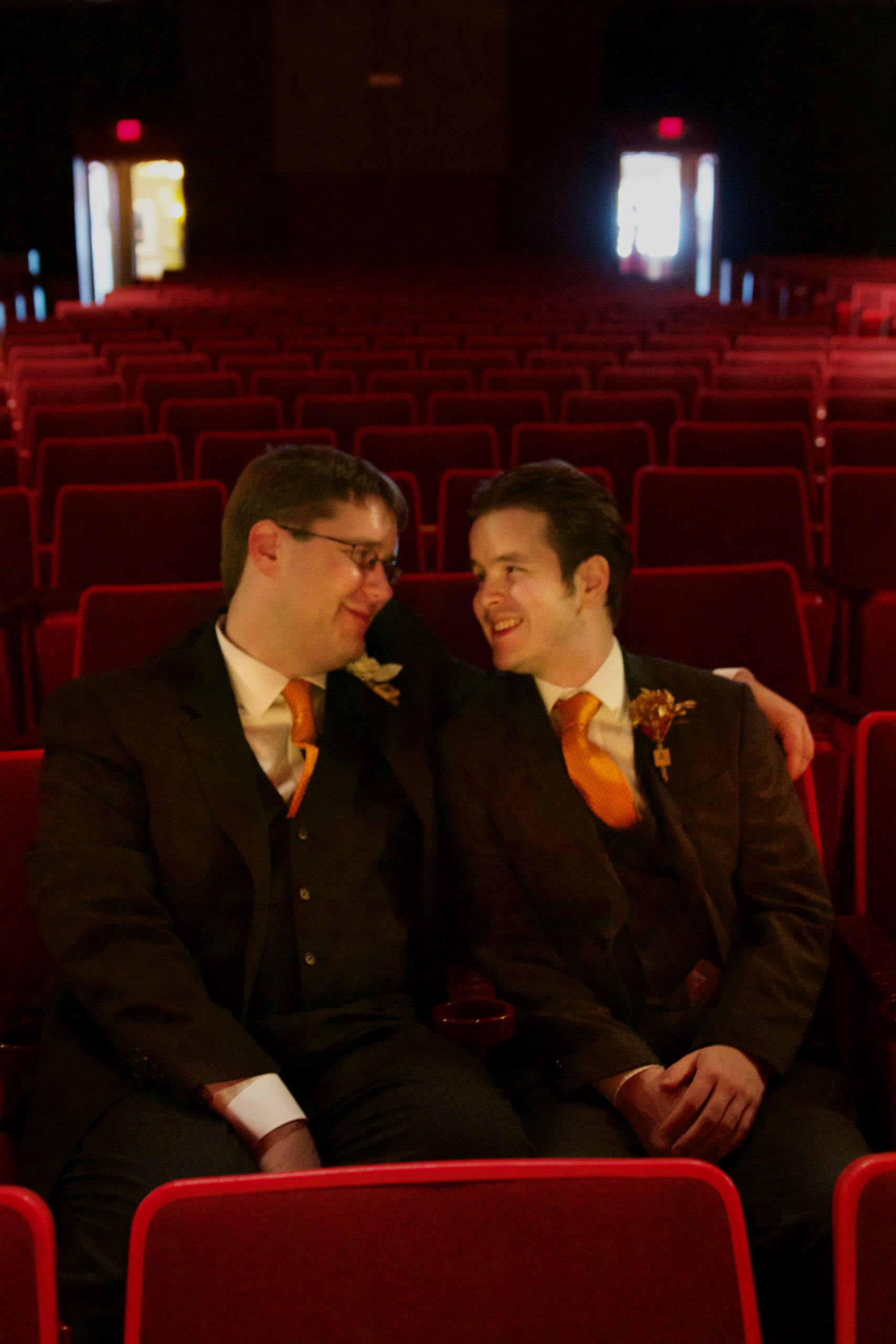 Aeon Affordable Homes Two men in suits with orange ties sit closely together, smiling, in an empty theater with red seats—it's part of Andrew's Story.