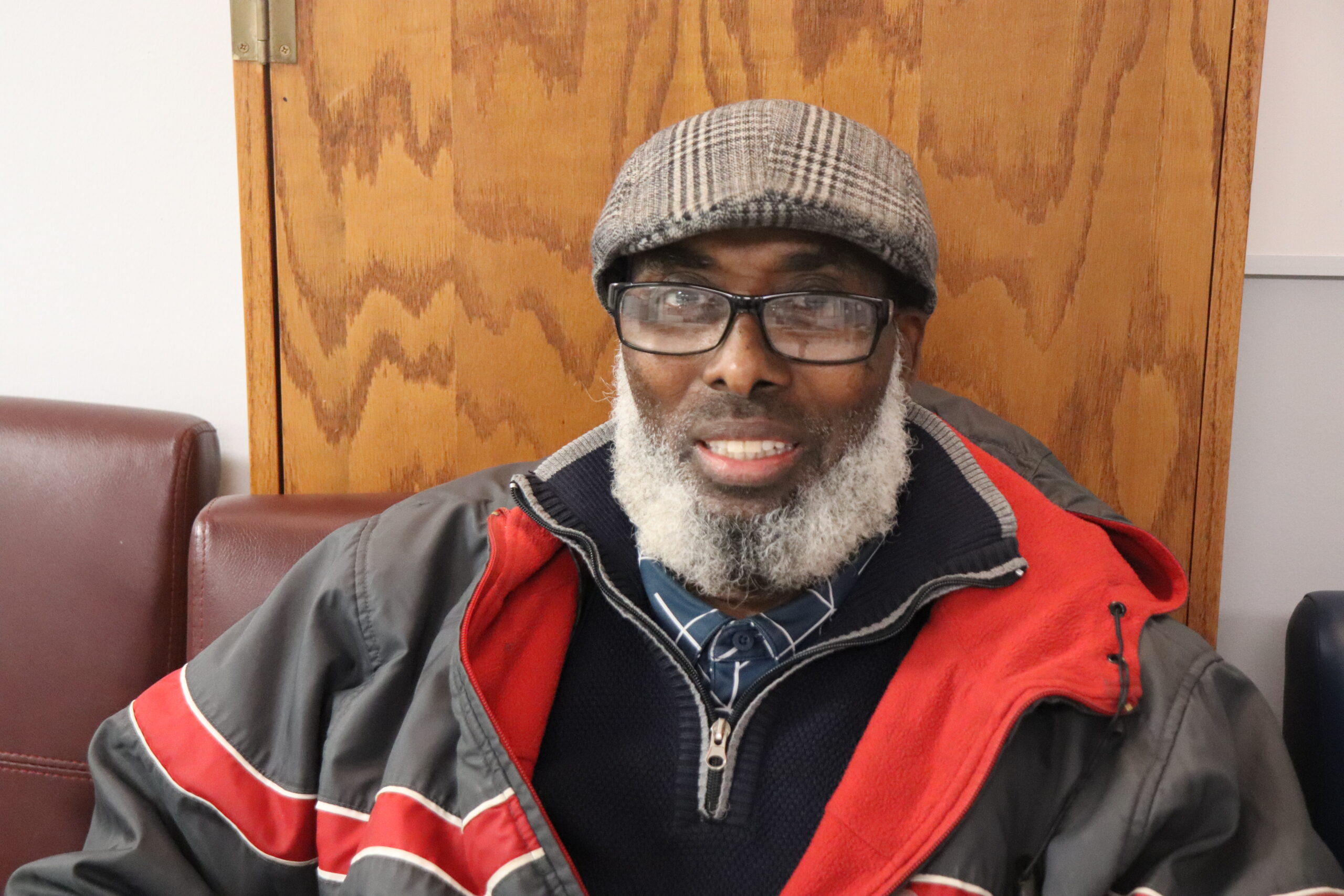 Aeon Affordable Homes A smiling man with a gray beard, glasses, and a flat cap sits on a maroon chair in a jacket, embodying Jama's Story.