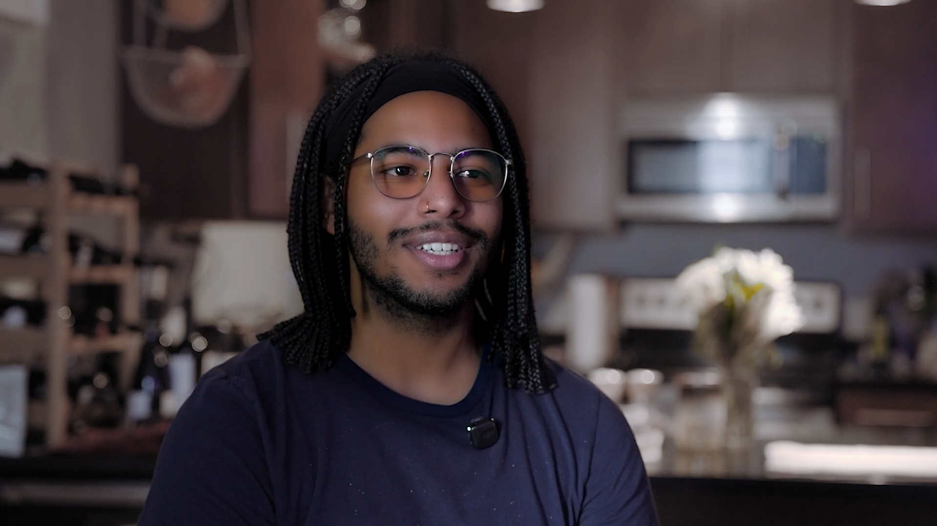 Aeon Affordable Homes A smiling person with glasses and braids sits in a kitchen, wearing a dark shirt and headset microphone.