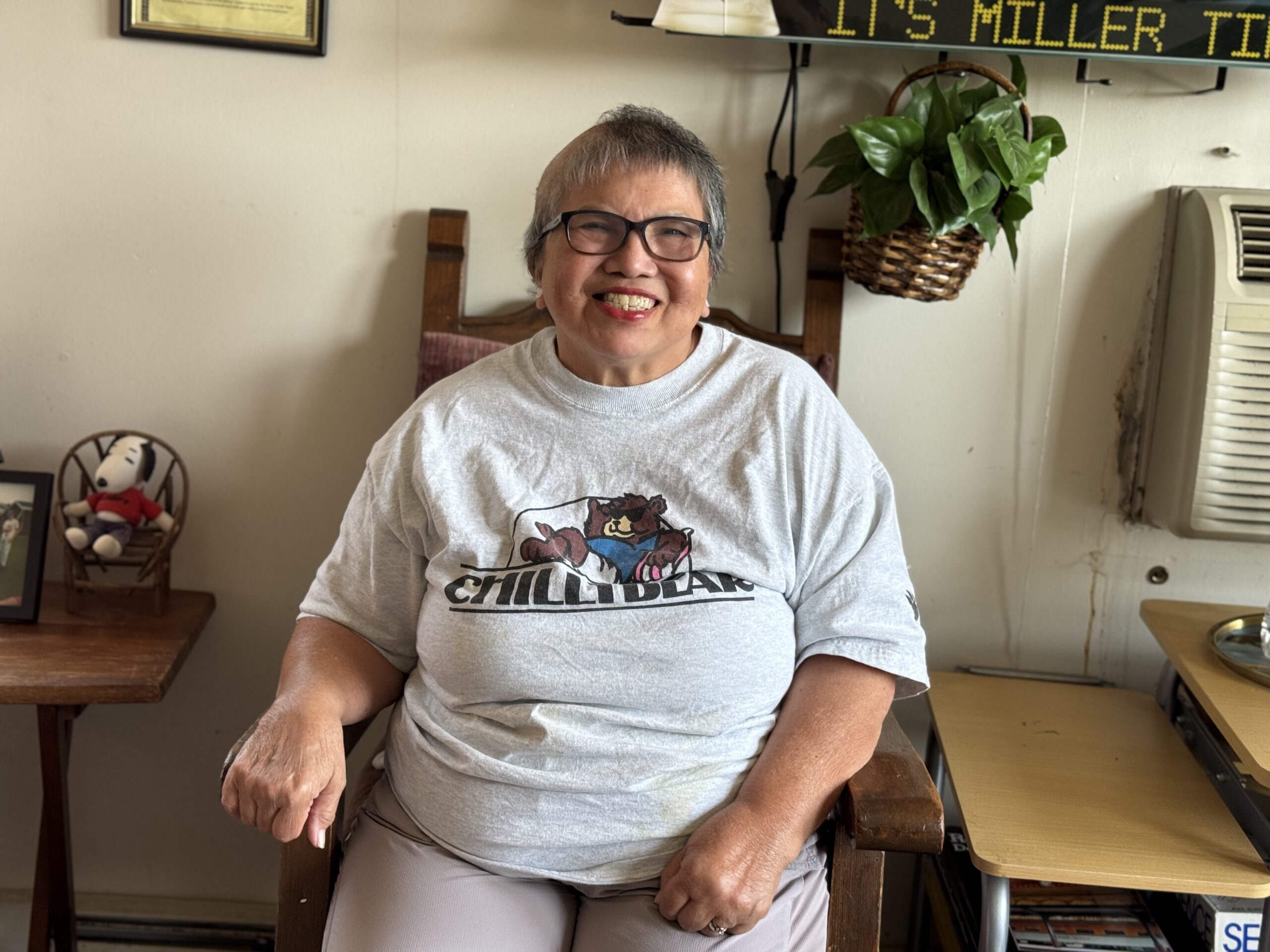 Aeon Affordable Homes Smiling woman with short gray hair sits indoors wearing glasses and a gray “CHILLI BEAR” T-shirt, sharing Amelia's Story.
